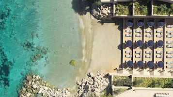 An aerial view of a beach resort with clear turquoise waters, a rocky shoreline, and a row of sunbeds with umbrellas.