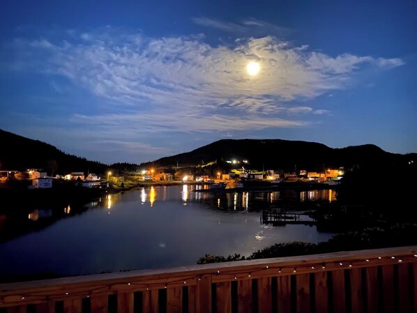 Une vue nocturne d’une ville au bord d’un lac, où les bâtiments se reflètent dans l’eau. Au premier plan, on aperçoit une terrasse en bois, et au loin, une colline.