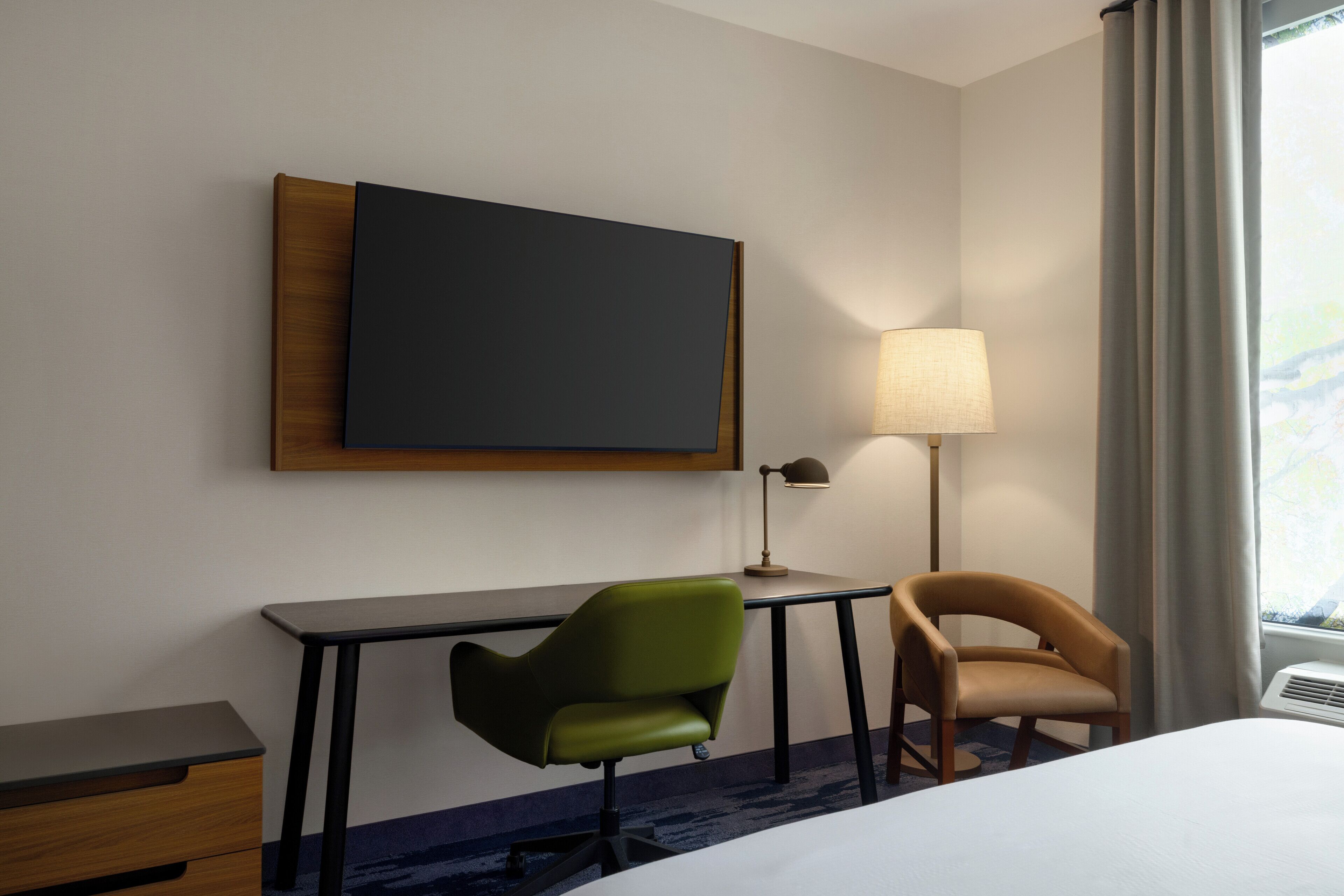 Une chambre d’hôtel avec un lit, un bureau, une chaise verte, une chaise marron, un téléviseur à écran plat fixé au mur, une lampe sur le bureau et une fenêtre avec des rideaux.