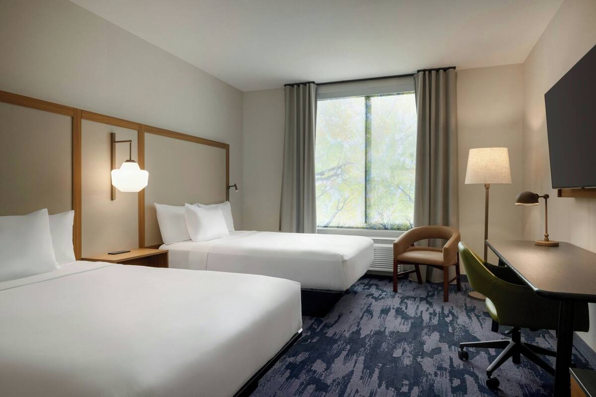 A hotel room with two beds, a desk, a chair, a TV, and a window with curtains.