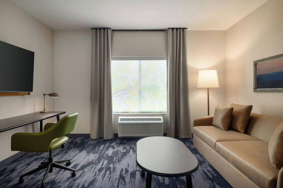 A hotel room with a desk, green chair, beige sofa, and a large window with curtains.