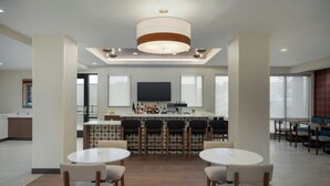 A modern hotel dining area with round tables and chairs, a central bar with stools, and a large TV screen. - Towneplace Suites By Marriott Lancaster, PA (Lancaster)