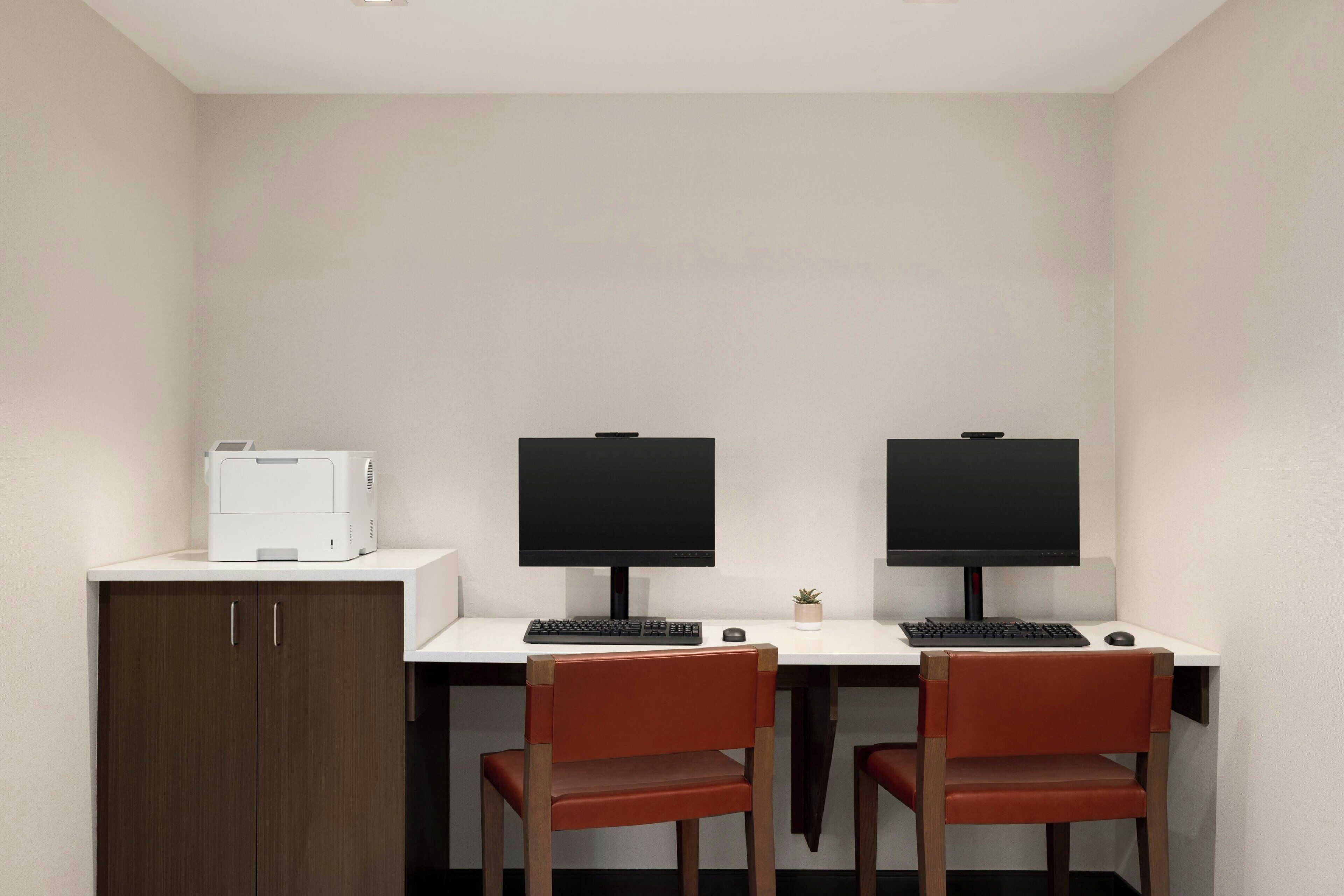 A room with two desks, each with a computer monitor, keyboard, and mouse. There is a printer on the left side of the room.