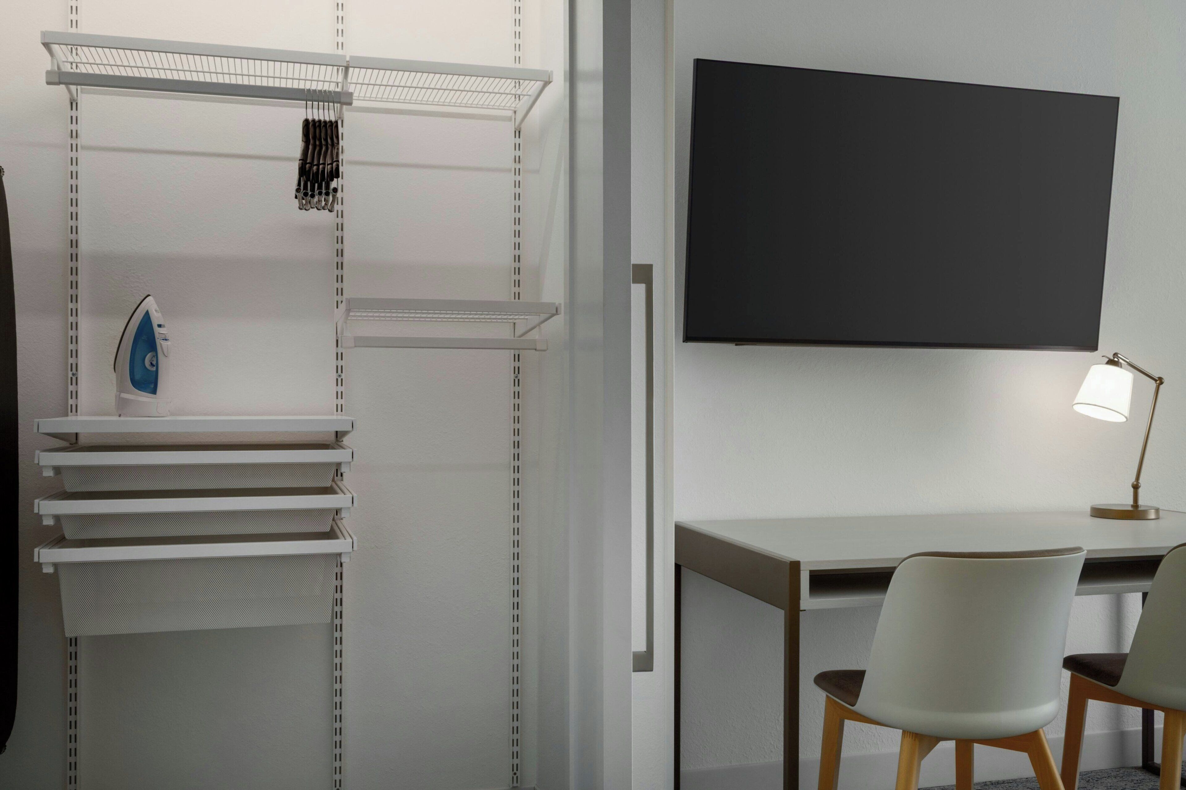A modern room with a desk, two chairs, a TV mounted on the wall, and a shelving unit with various items.
