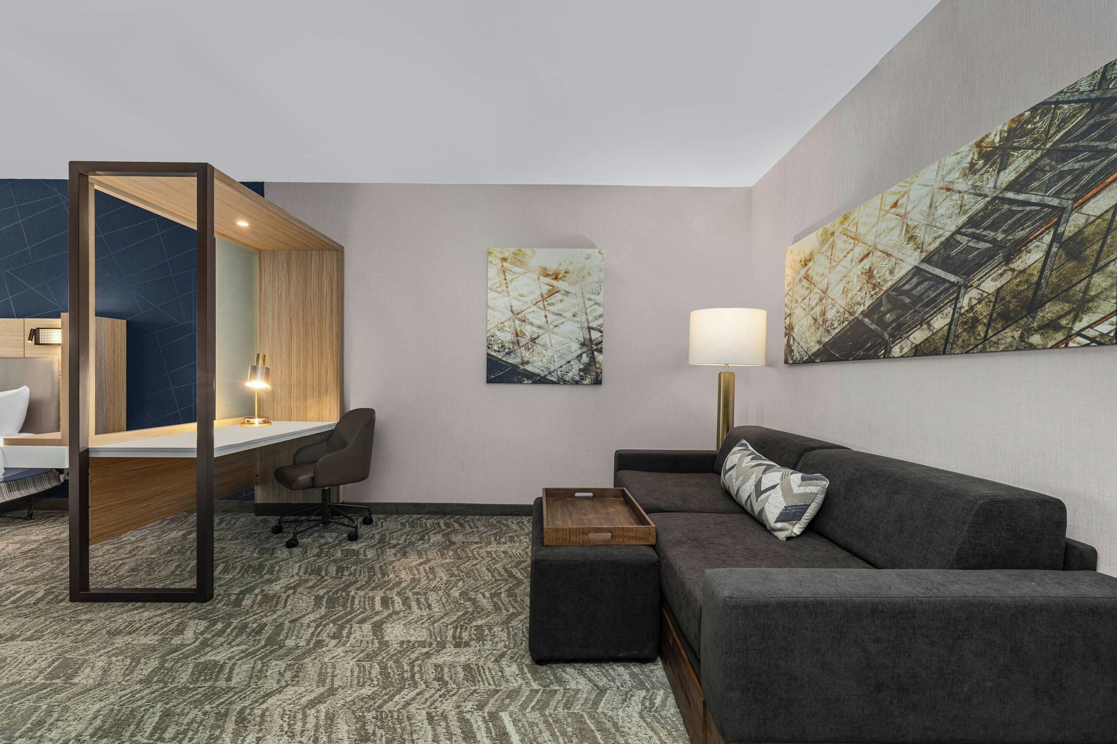 Una habitación de hotel moderna con escritorio, sofá, una mesita y una silla. Hay dos cuadros en la pared y una lámpara en la mesita de noche.