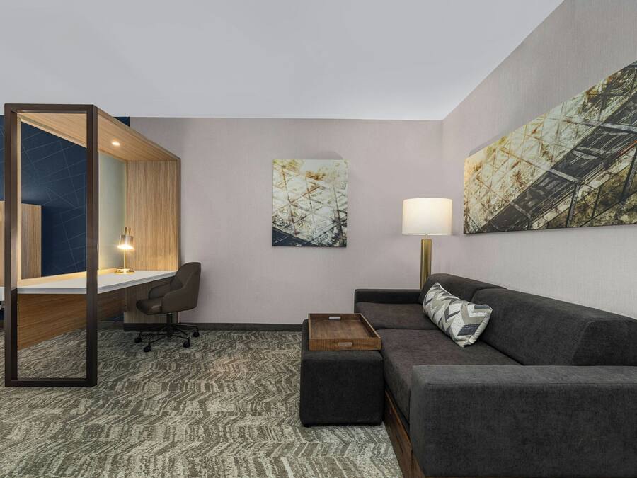 Una habitación de hotel moderna con escritorio, sofá, una mesita y una silla. Hay dos cuadros en la pared y una lámpara en la mesita de noche.
