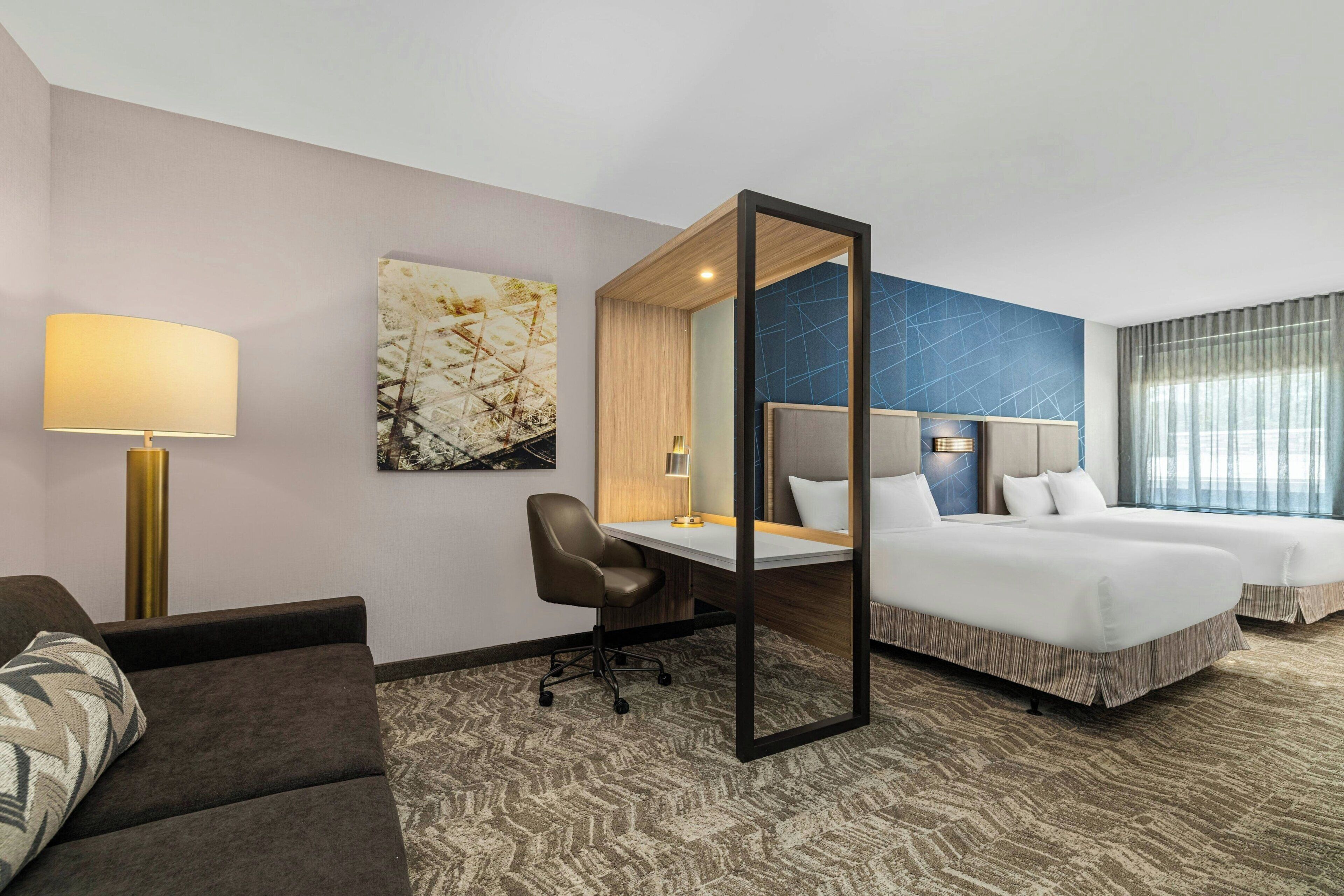 Une chambre d’hôtel équipée d’un bureau, de deux lits, d’un canapé, d’une lampe et d’œuvres d’art accrochées aux murs.
