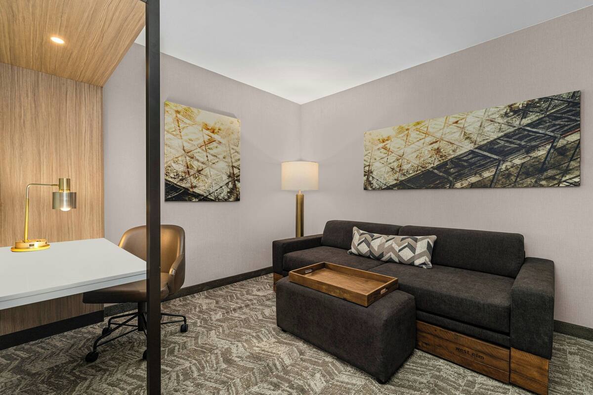 A modern room with a desk, office chair, sofa, and ottoman. There are two abstract paintings on the walls and a floor lamp.