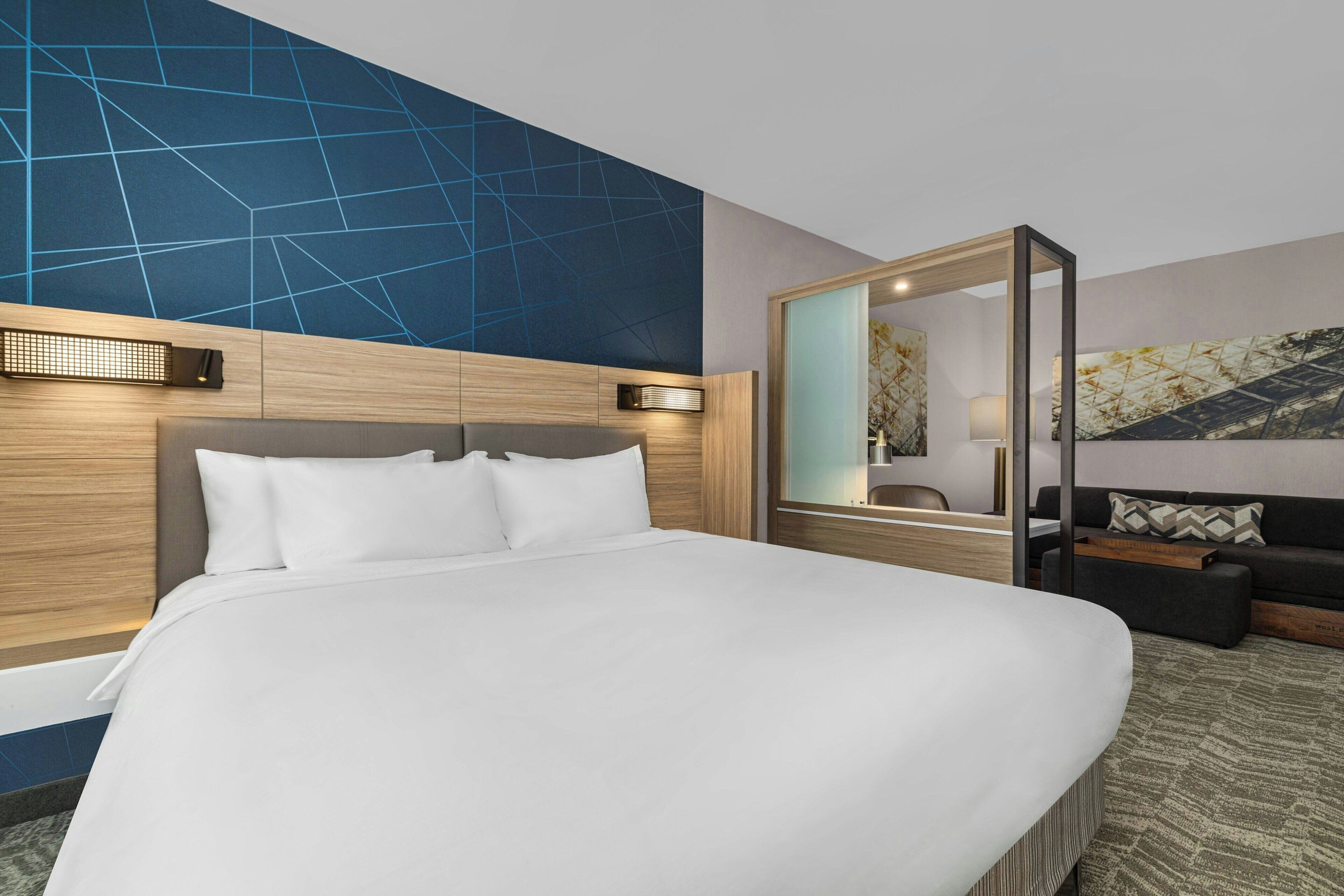Habitación de hotel moderna con una cama grande, cabecero de madera, un espejo, un sofá y una pared con un diseño geométrico.
