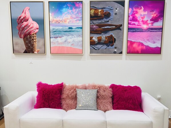 Un sofá blanco con almohadones rosados y plateados de textura suave, y tres cuadros colgados encima.
