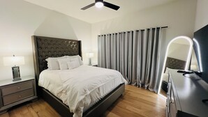 A hotel room with a large bed, a ceiling fan, a nightstand with a lamp, a dresser with a mirror, and a window with curtains. - Perfect getaway in Corktown! (Detroit)