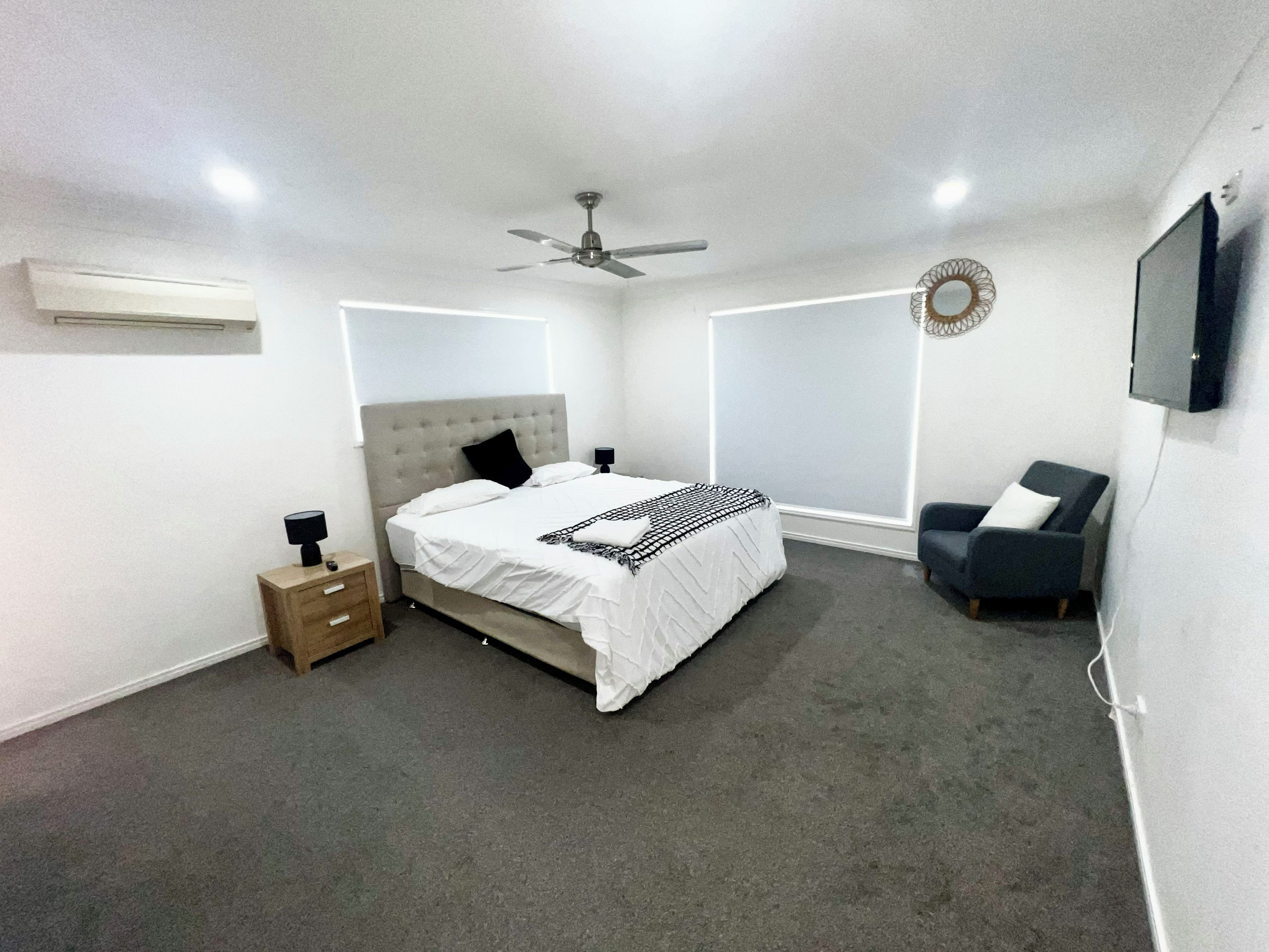 Un dormitorio con cama, mesitas de noche, una silla, ventilador de techo, televisor y ventana.