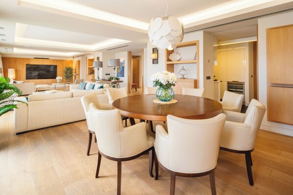 Een moderne woonkamer met een ronde eettafel, witte leren stoelen, een grote flatscreen televisie en een houten kast.