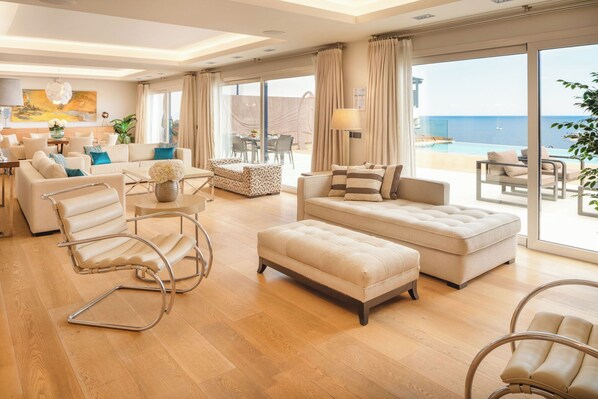 Amplio salón con un ventanal grande que da a una piscina y al mar. Hay un sofá, una silla y una mesa con una lámpara.
