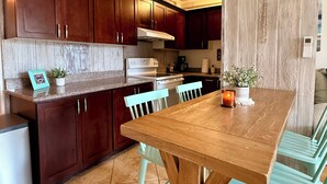A kitchen with dark wooden cabinets, a wooden dining table, and teal chairs. - Kaua Corner (Paia)