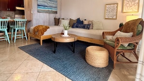 A living room with a sofa, coffee table, and wicker chair. There is a dining area with a table and chairs. - Kaua Corner (Paia)