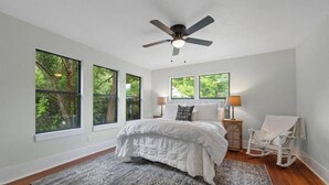 Un dormitorio con cama, ventilador de techo, mesita de noche, una silla y ventanas grandes con vistas a árboles.