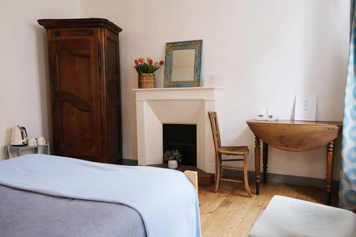Une chambre à coucher comprenant un lit, une armoire en bois, une cheminée, une chaise, une petite table et un miroir.