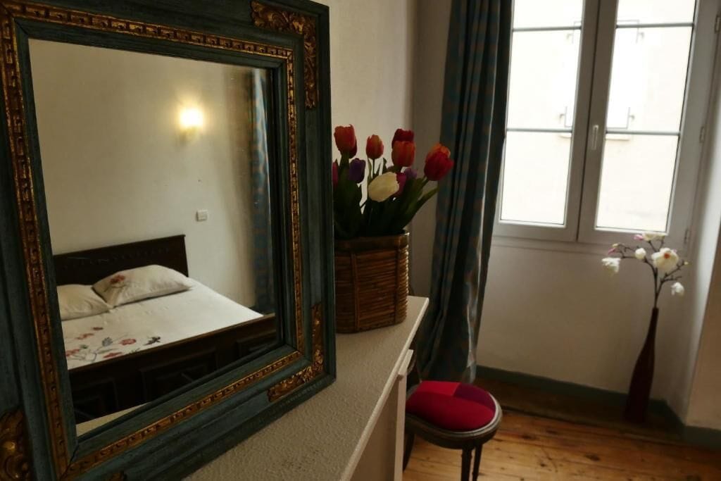 Un dormitori amb un mirall gran, un llit amb roba de llit floral, un vas amb flors, una cadira i una finestra amb cortines.