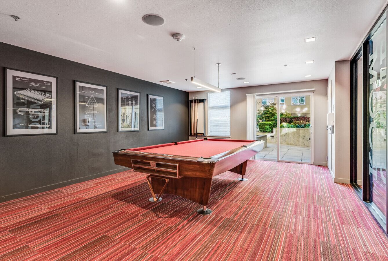 A pool table in a room with grey walls and a red carpet. There are framed pictures on the wall and a door leading outside.