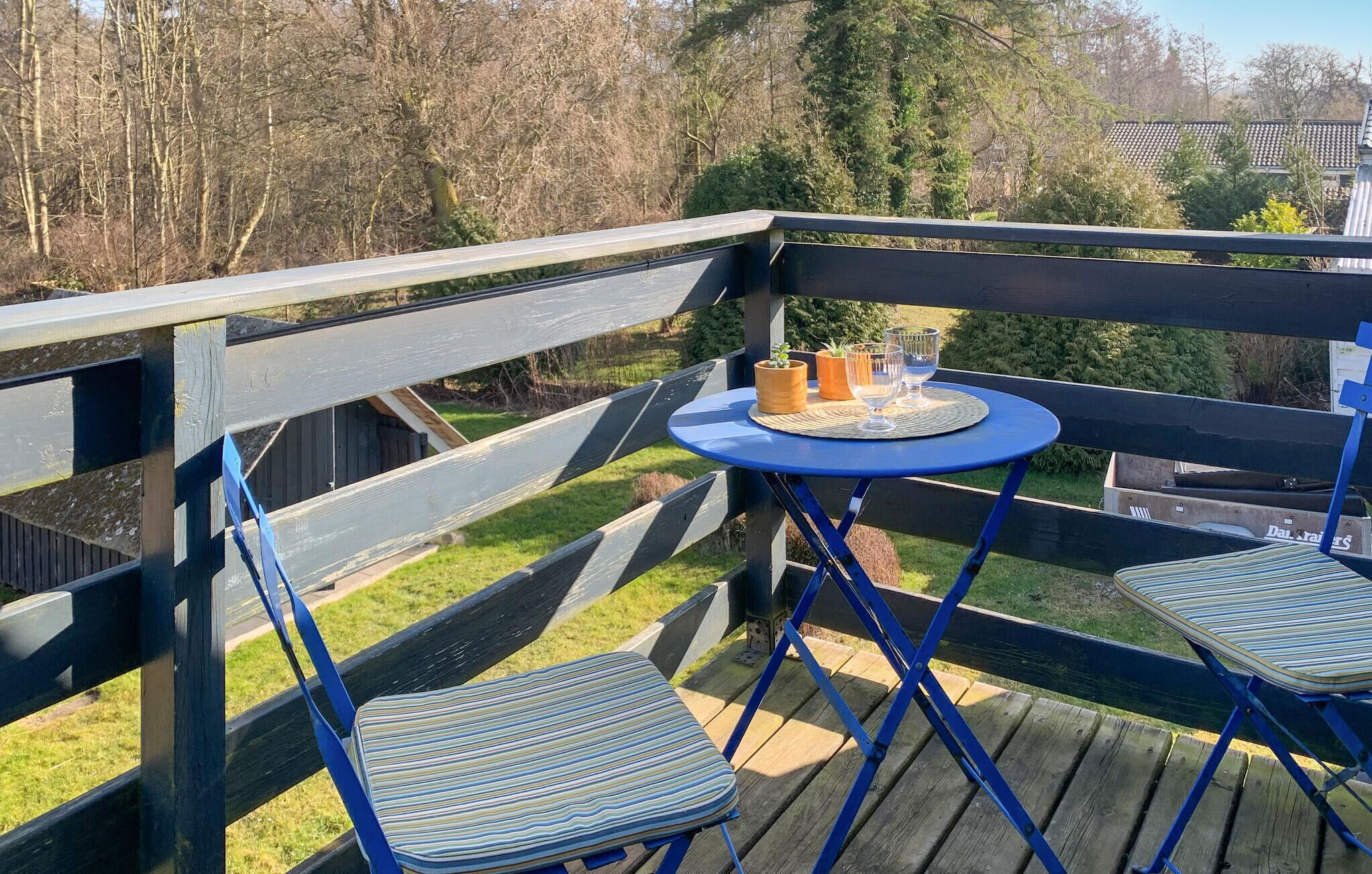 Una terraza con una mesa azul y dos sillas a rayas, una pequeña planta en maceta y un juego de vasos sobre la mesa.