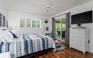 Une chambre à coucher avec un lit, un ventilateur de plafond, une télévision, une porte-fenêtre coulissante et une commode blanche.