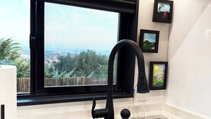A modern kitchen with a black faucet, a wooden cutting board, a window with a view of greenery, and framed pictures on the wall. - ArtNest: Full Kitchen & bath, Dishwasher, W/D, Firepit, Private, Parking, Safe (Escondido)