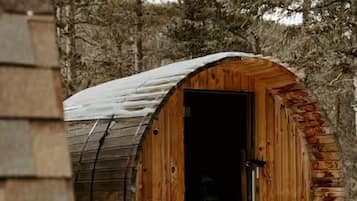 Una cabaña de madera rústica con un gran ventanal y dos ventanas pequeñas, rodeada de nieve y árboles.