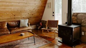 A cozy room with a wooden ceiling, a brown leather sofa, a wooden coffee table, a stone fireplace, and a window with a view of trees.