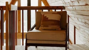A cozy attic room with a wooden sloped ceiling, a single window, and a cushioned chair with a patterned pillow.