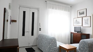 Ein Wohnzimmer mit Sofa, einem kleinen Tisch, einem Fernseher auf einem Schrank und einer Tür mit Fenster.