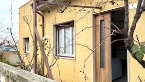 Ein gelbes Gebäude mit einer Holztür, einem Fenster mit Rollläden und einem Balkon mit Steinbalustrade.