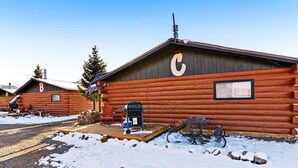 Eine Holzblockhütte mit einem großen 'C' an der Seite, einem Grill davor und Schnee auf dem Boden.