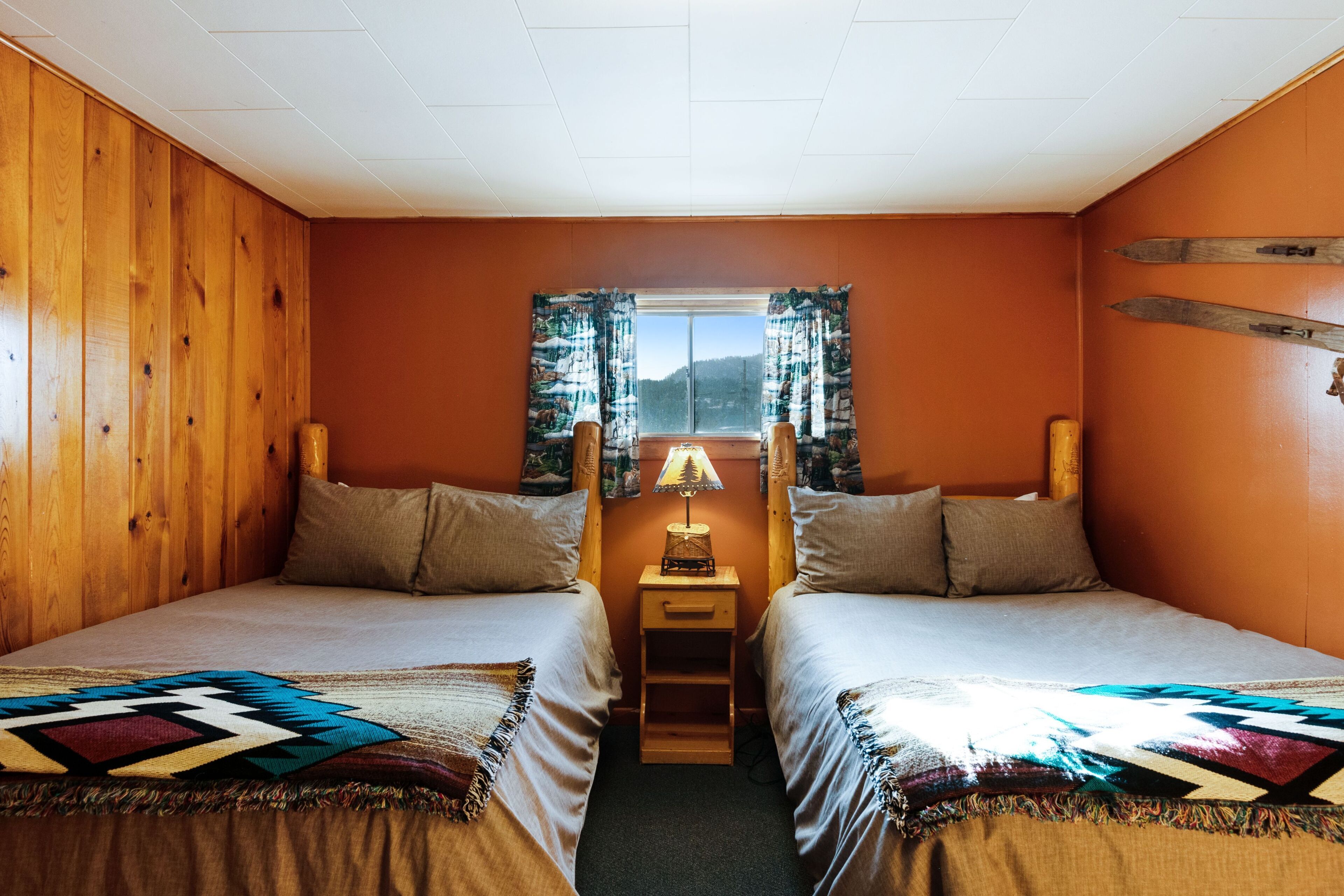 Habitación con dos camas, paredes de madera, ventana con cortinas, mesita de noche con lámpara y vista a la montaña.