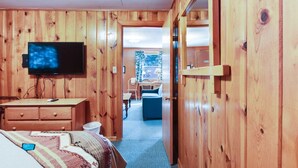 Ein Zimmer mit Holzwänden, einem Bett, einem Schreibtisch, einem Fernseher und einer Tür zu einem weiteren Raum.