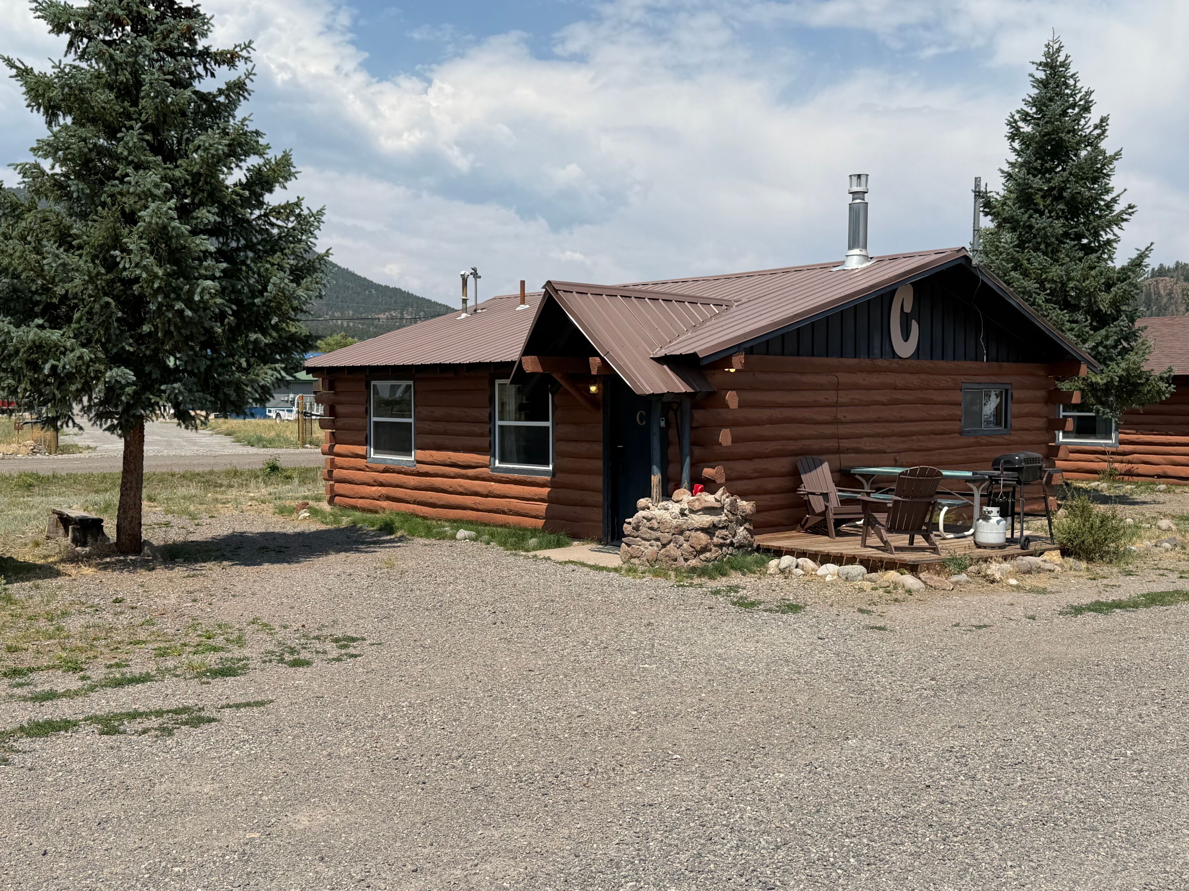 A log cabin with a gravel driveway, outdoor seating, and a mountain backdrop.