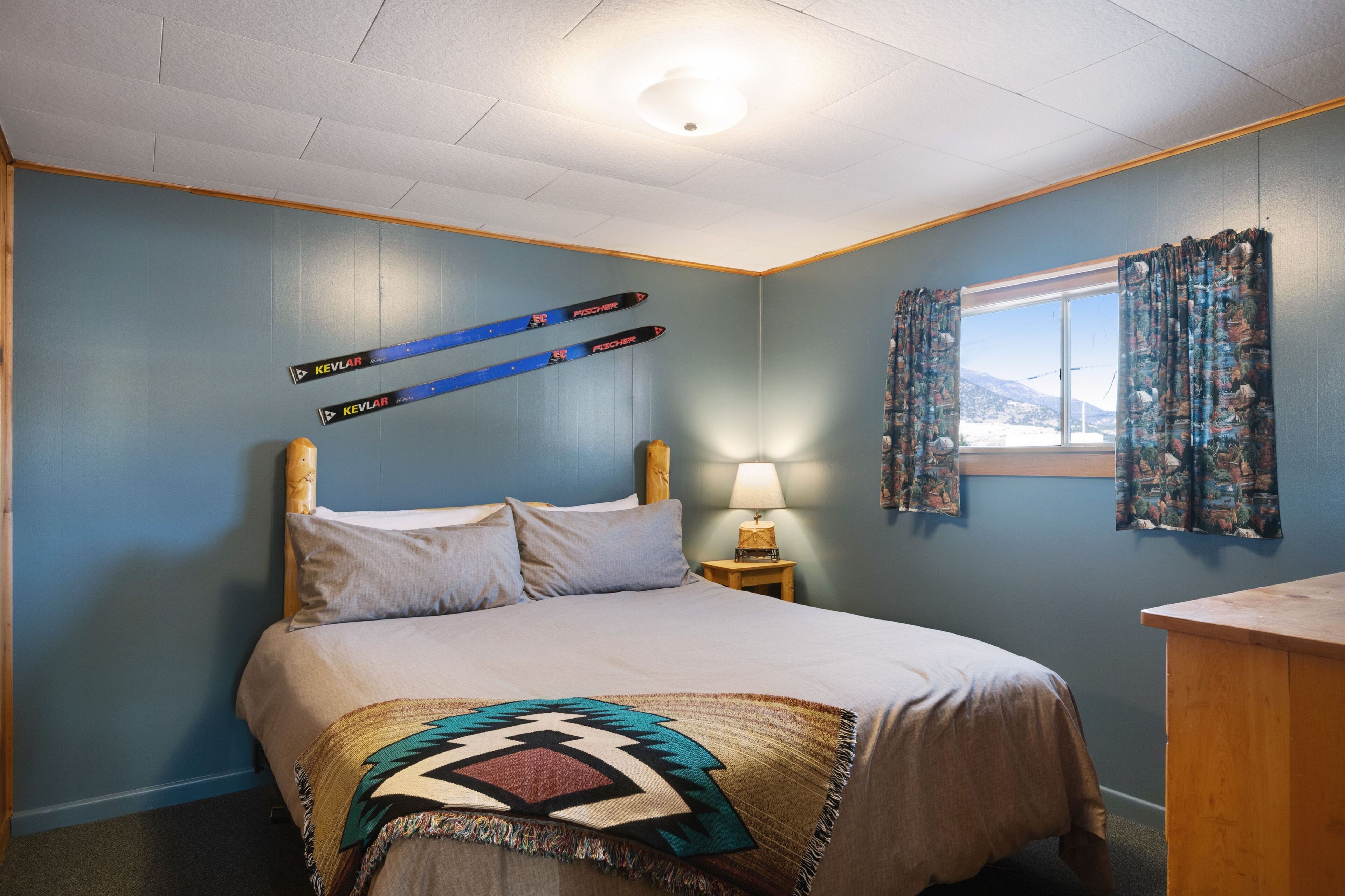 Een slaapkamer met een bed, nachtkastje met lamp, raam met gordijnen en ski’s aan de muur.