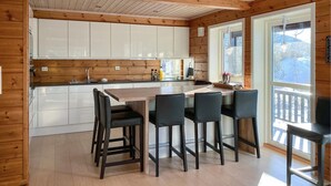 A modern kitchen with wooden paneling, white cabinets, a central island with bar stools, and large windows offering a view of the outdoors. - Cozy home in Stryn with sauna (Stryn)