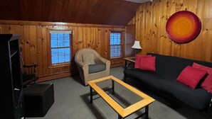 Una sala de estar acogedora con un ventilador de techo, un sofá negro con cojines rojos, una mesa de centro de madera, un sillón beige y un televisor sobre un mueble.