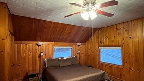 Un dormitorio con techo de paneles de madera, ventilador de techo, dos ventanas, una cama y una mesita de noche.