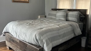 A bedroom with a wooden bed frame, a white comforter, and a gray striped pillow. There is a nightstand with a lamp, a painting on the wall, and a window with sheer curtains. - Historic 3 Bedroom home, steps away from the Capitol, Clay Center and Ballpark. (Charleston)