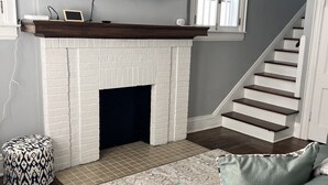 A living room with a white brick fireplace, a mounted TV, a staircase, and a patterned rug. - Historic 3 Bedroom home, steps away from the Capitol, Clay Center and Ballpark. (Charleston)