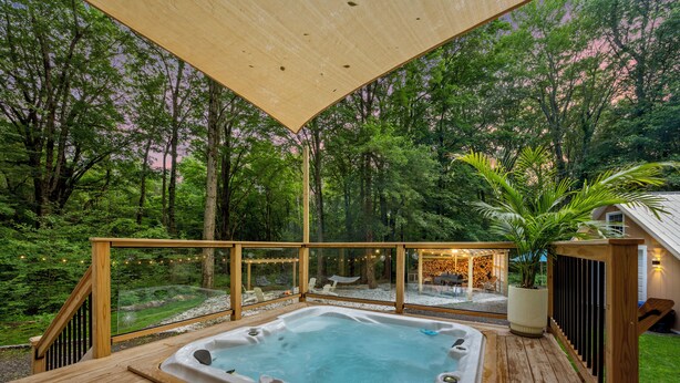Un jacuzzi en una terraza de madera con barandas de vidrio, rodeado de un área boscosa.