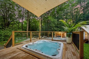 A hot tub on a wooden deck with glass railings, surrounded by a forested area.