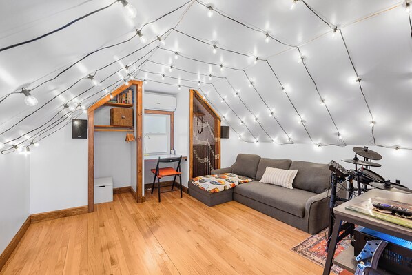 A cozy attic room with a wooden floor, a gray sofa, a desk with a chair, and a drum set.