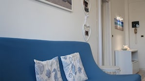 A blue sofa with decorative pillows, a white wall with framed photos, and a small white table with a lamp. - P051 – Porto Recanati, comfortable two-room apartment with sea view (Porto Recanati)