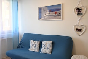 Ein Zimmer mit einem blauen Sofa, zwei gemusterten Kissen, einem gerahmten Gemälde und herzförmigen FotoRahmen an der Wand.