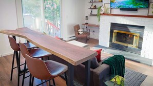 A modern living room with a wooden dining table, leather chairs, a fireplace, and a large TV mounted above it.