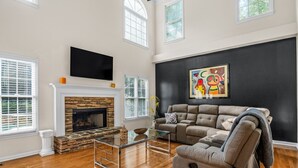 A spacious living room with a fireplace, a large sofa, and a flat-screen TV mounted on the wall. - New! Lux Mansion, 5 BR 4 BA, Heated Pool, Sleeps 12+ (Fayetteville)