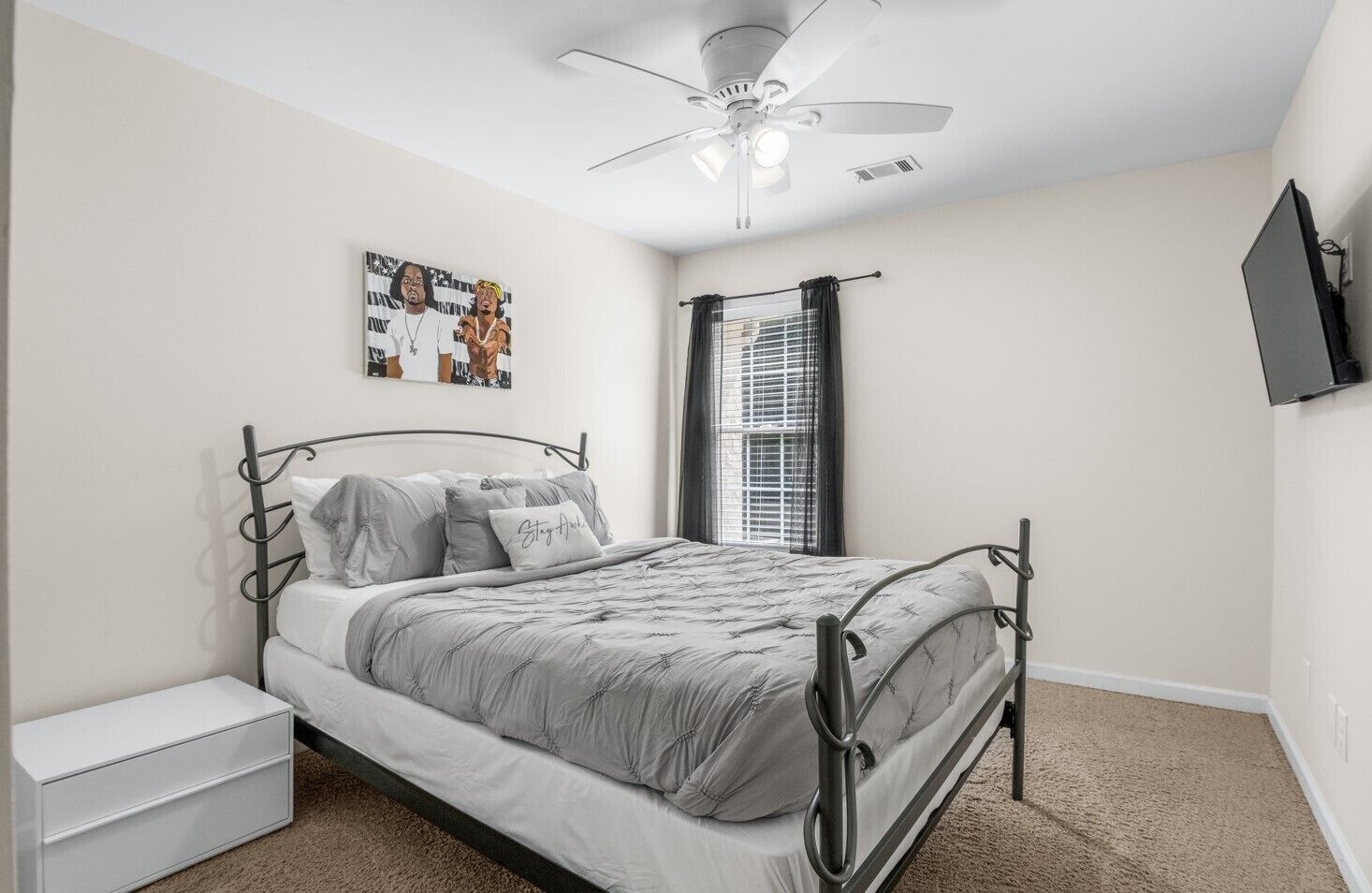 A bedroom with a bed, a nightstand, a ceiling fan, a window with curtains, and a wall-mounted TV.
